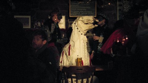 A still from Pen Mari: a white person-sized puppet with a horse's skull perched on the top. Sat amongst a busy, dimly-lit pub.