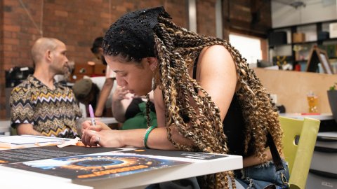 A young woman with long braids leans over a studio table, writing on a large printed sheet as others gather nearby in a lively, creative space.