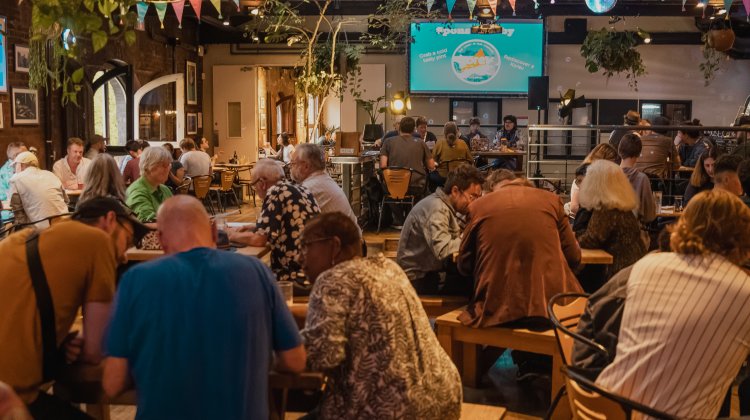 A vibrant Café & Bar scene with patrons engaged at wooden tables, enjoying drinks and a lively atmosphere, under colourful decorations and plants.