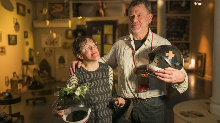 A man and woman (Andrew and Eden Kötting) stand in a warmly lit room, embracing. The woman holds a helmet with plants; the man holds a helmet with a band-aid. They appear content.