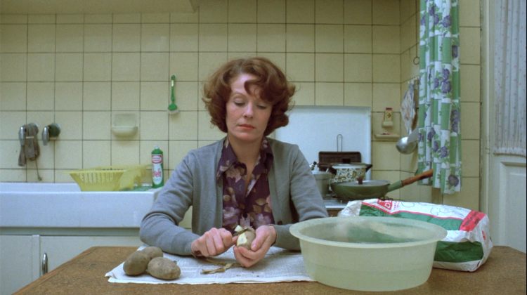 A photograph of a woman sat at a kitchen table, peeling potatoes and looking downwards against the background of a simple seventies kitchen.