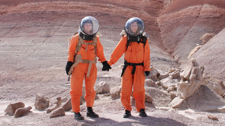 Two women in spacesuits in a barren landscape holding hands
