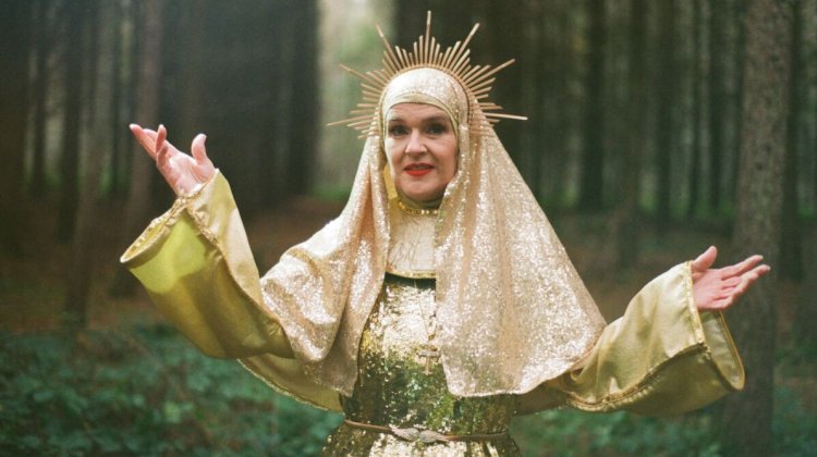 A figure in a golden, shimmering robe with a halo stands in a forest, arms raised and surrounded by tall trees.