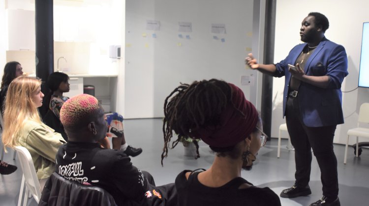 A speaker in a blue jacket engages with an audience seated in a modern room, featuring a screen displaying images.