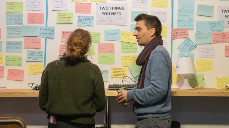 Two people standing in front of a board of multi coloured post-it notes in discussion