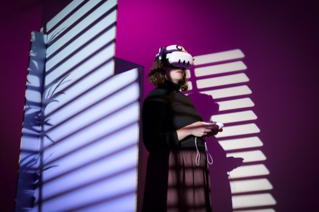 A woman stands with a VR headset on and controllers in here hand, the background is purple and pink with various shadows.