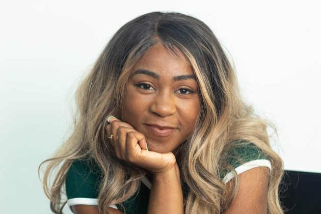A young Black woman sits with a hand under her chin, she has long blonde hair and smiles softly into the camera.