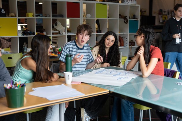 A group of diverse young adults collaborates around a table with notes and materials, engaged in discussion in a creative workspace.