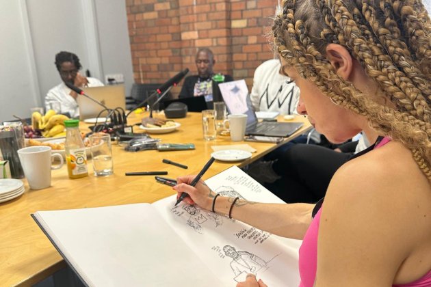 A person with braided hair sketches in a large notebook during a meeting at a wooden table with food and drinks present.