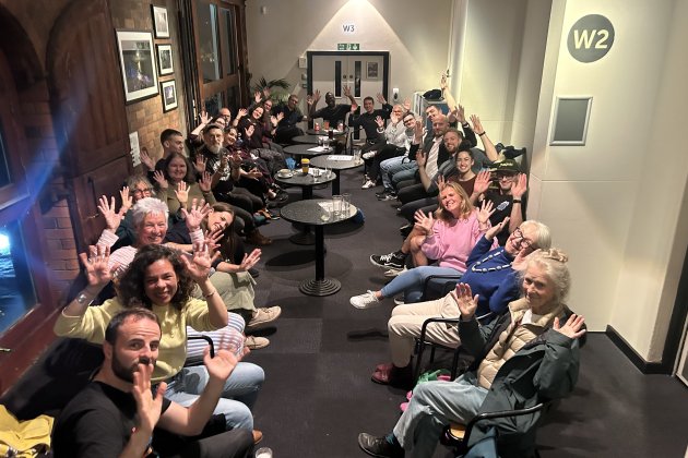hoto of Deaf Conversations About Cinema discussion group at Watershed. A large group of people sitting in a long corridor together - waving their hands in the air after the discussion.