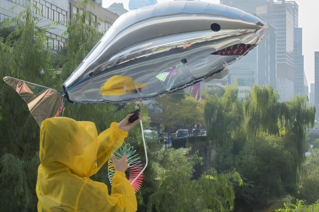 Person in a yellow raincoat holding a large silver shark-shaped balloon near a river with trees and city buildings in the background.