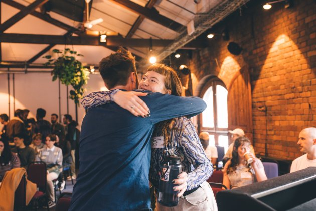 Two people hug and smile in a room with exposed brick walls, while others sit and watch in the background.