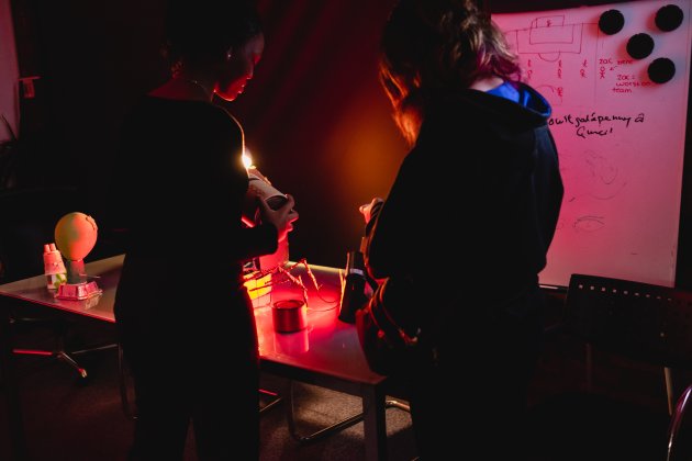 Two people examine a small object at a cluttered worktable under red light, beside a whiteboard covered in sketches and notes; the mood is focused and tense.
