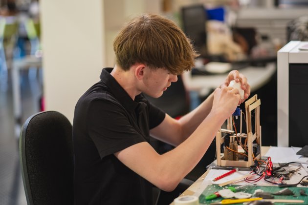 A focused young man in a black polo carefully assembles an electronics project at a workbench, surrounded by wires and tools in a busy workshop.