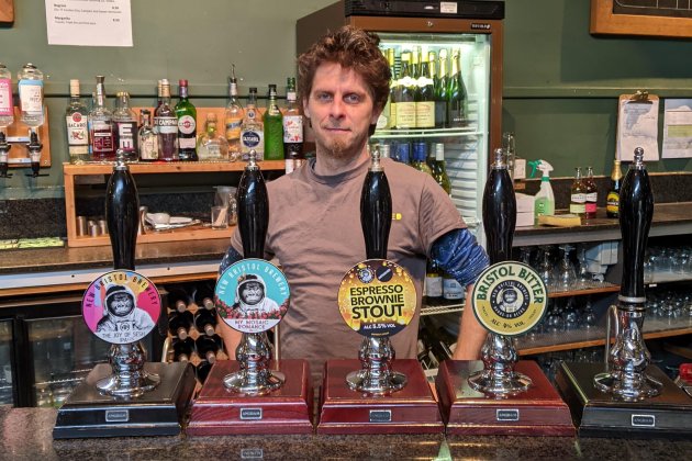 An image of a man standing behind four beer pumps.