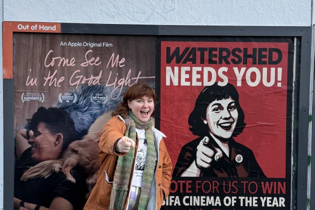 An outdoor poster site - a person stands to the side smiling and pointing in the same pose as the person in the poster which also has the words' Watershed Nees