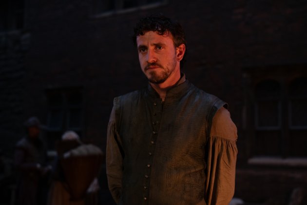 A young man stands in dim light, wearing a historical tunic with an embroidered collar, while figures gather in the background.