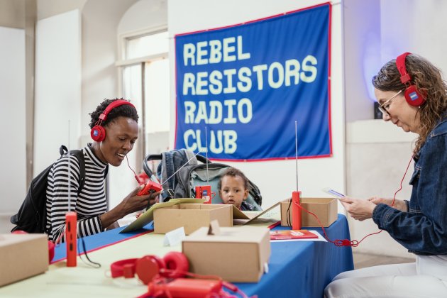 A young Black women sits smiling with red headphones on and a banner above her reads Rebel Resistors Radio Club. 