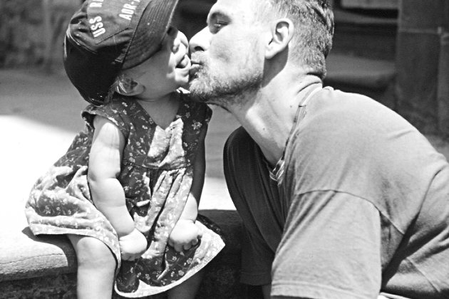 A black and white photo of the artist Andrew Kötting kissing his daughter Eden on the cheek.