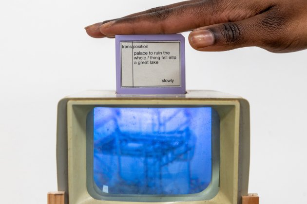 A hand hovers over a small screen atop a retro TV showing a blue image. The screen displays text with words about a "palace" and a "lake." The vibe is artistic and thought-provoking.