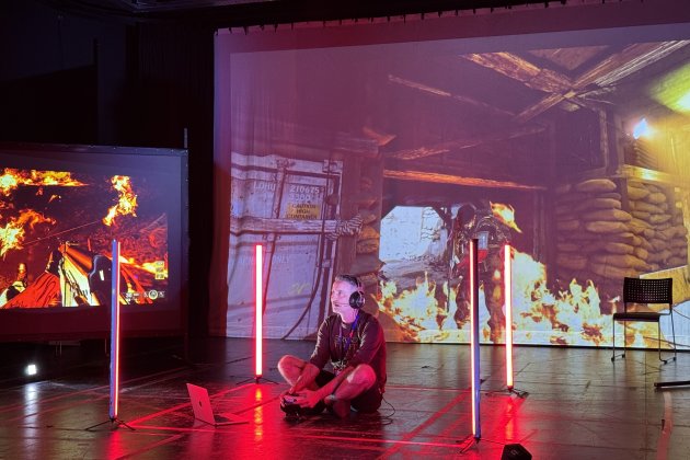 A person sits cross-legged on the floor, wearing headphones, surrounded by vertical red lights. Intense video game scenes with fire project in the background.
