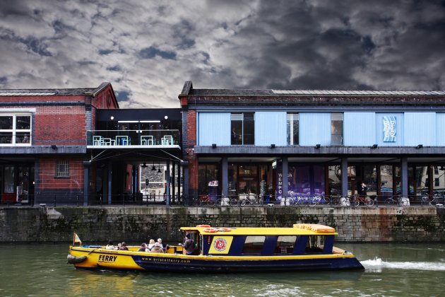 exterior shot of the Watershed building, including the balcony, there is a yellow ferry on the water passing the building and the sky is dark and stormy.