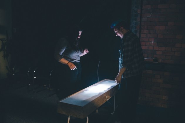 Two people stand in a dimly lit room, focused on a glowing rectangular table. The scene conveys curiosity and concentration against a brick wall backdrop.