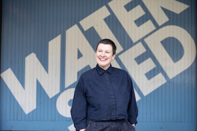 A smiling woman stands outside the Watershed blue panel - she has short cropped hair, and is look away from the camera.