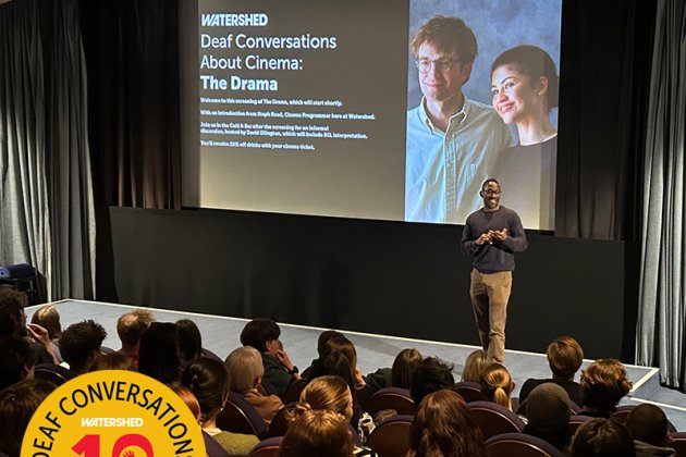 Inside a full cinema, man at the front of the audience introduces a Descriptive Subtitled screening of The Drama