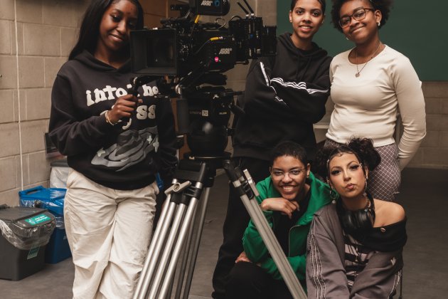 Five young people posing with a professional ARRI camera on a tripod in an indoor setting with concrete walls.