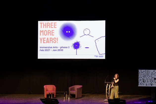 Speaker stands on a dimly lit stage beside a slide reading “THREE MORE YEARS!” with colorful drawings, creating an optimistic, creative tone.