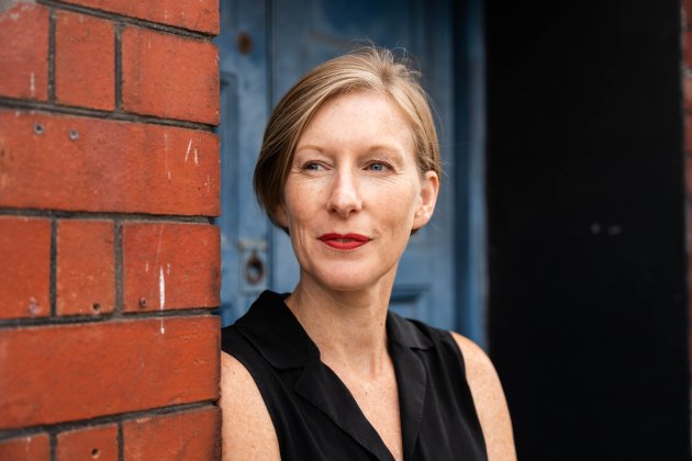 A woman with short hair leans against a brick wall with a blue background, she is smiling and looking to here right.