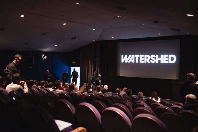 Audience gathering in a cinema with purple seating, facing a large screen displaying the word "WATERSHED."