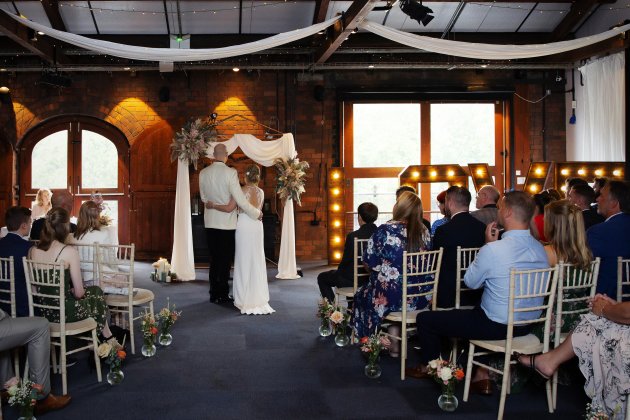 A couple stands under a floral arch at their wedding ceremony, surrounded by guests seated in chairs, with decorative lights in the background.