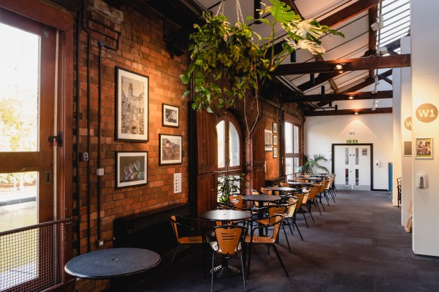 A spacious, warmly lit café with brick walls, framed art, and black tables lined up beside large windows letting in natural light.