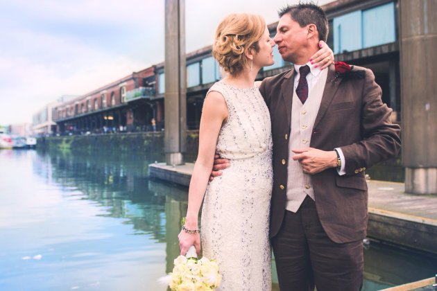 A bride in a sparkly white dress holds a bouquet while embracing her partner by a serene waterfront, surrounded by a scenic backdrop.