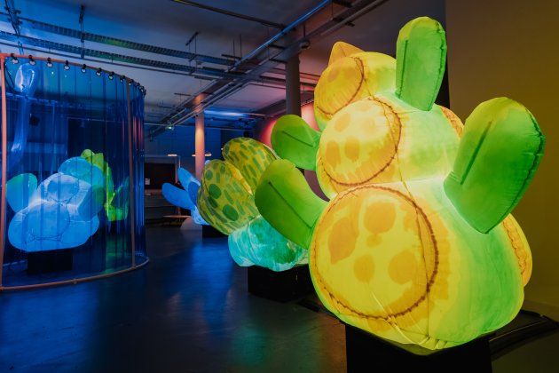 Colorful illuminated inflatable flower sculptures displayed in a modern gallery, with blue reflections in a cylindrical enclosure.