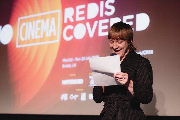 A person in front of a screen speaks to an audience form their notes