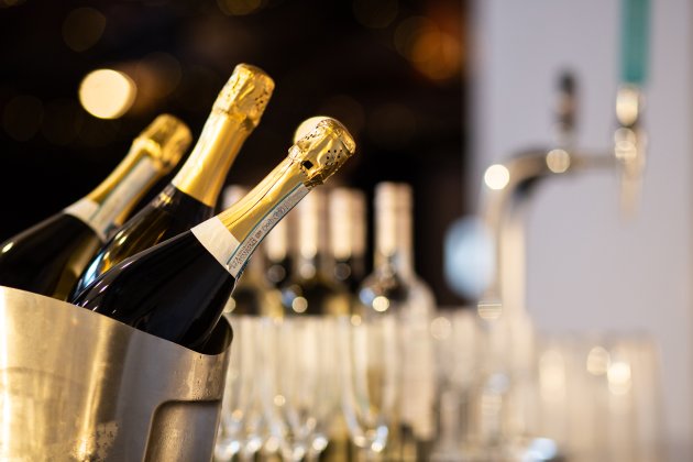 A metal bucket filled with champagne bottles stands near a row of wine bottles and glasses, set against a softly lit background.