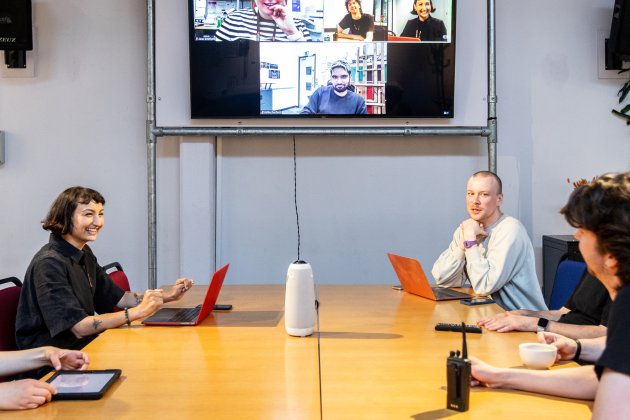 A group of people in a conference room engaged in a virtual meeting, with laptops and a screen displaying remote participants.