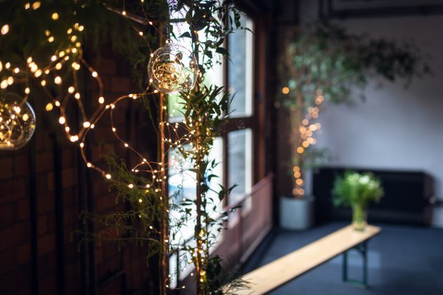 Cozy indoor space adorned with fairy lights and greenery, featuring a wooden bench and large windows letting in natural light.