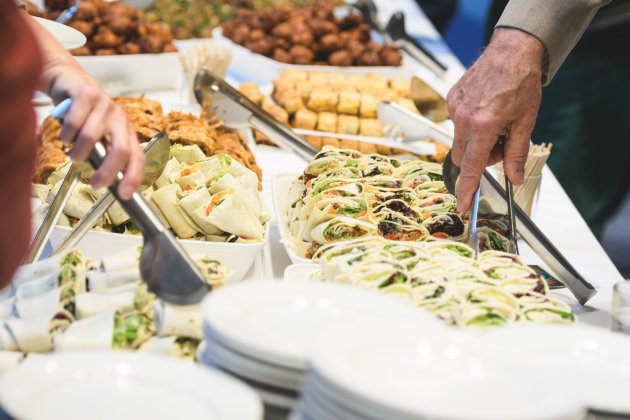 A buffet display featuring various wraps and appetizers, with hands serving from trays and plates in the foreground.