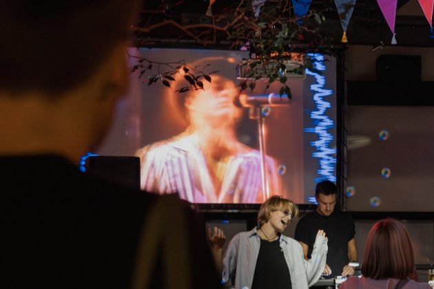 A lively scene featuring a crowd watching a musician perform on a screen, with colorful decorations and a DJ setup in the background.