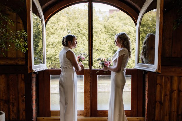 Two elegantly dressed individuals stand by a large window, holding floral bouquets and looking out at a lush, green landscape.