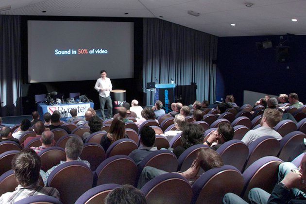 A speaker presents to an audience in a cinema setting, with a slide displaying "Sound in 50% of video" on the screen behind.