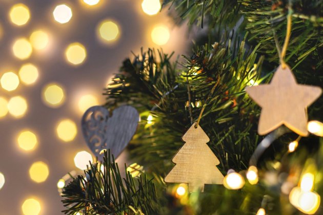 Close-up of a Christmas tree with wooden ornaments, including a star and a tree, against a blurred backdrop of warm, glowing lights.