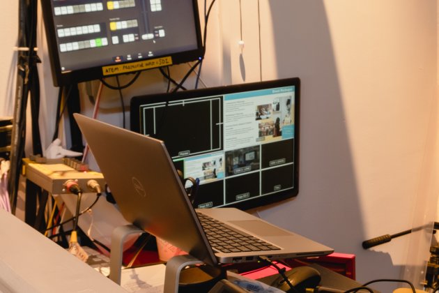 A laptop and two monitors on a production desk, displaying video feeds and control options for a live event setup.