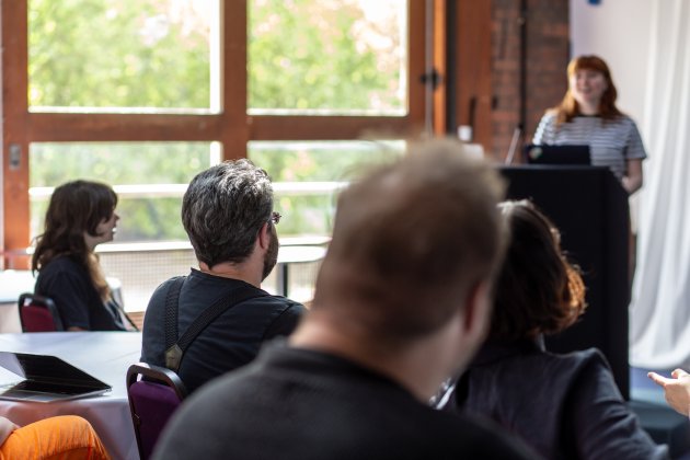 A group of people seated in a conference room, attentively listening to a speaker at a podium, with large windows in the background.
