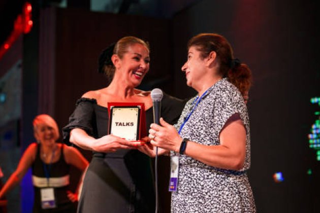  Two women on stage, one holding a red award plaque labeled 'TALKS,' while speaking into a microphone. Colorful lights in the background.