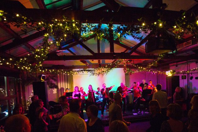 Indoor live band performance in the Waterside 3 event space at Watershed, with colourful stage lights, decorated ceiling beams, and an audience dancing and socialising.